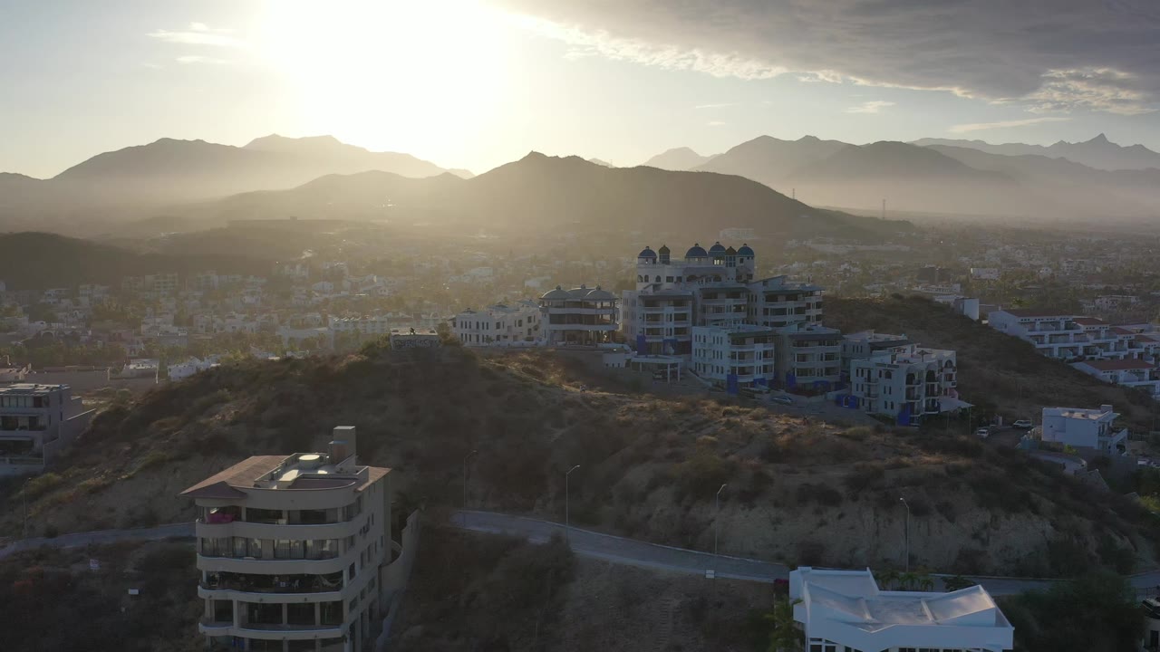 Los Cabos aerial frame showing apartment-style buildings on a dry ridge, scattered homes in the valley, strong sun haze, layered mountains, and no visible people.