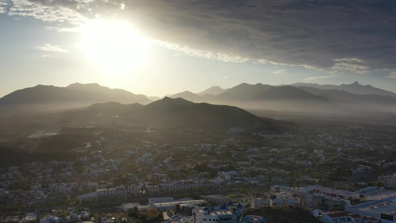 Opening frame of Los Cabos, Mexico, showing bright sun above layered mountains, a dark foreground hill, rooftops below, and no visible people.