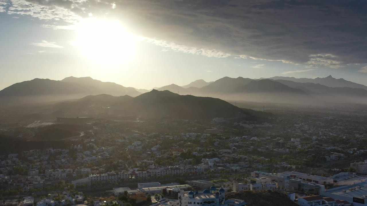 Los Cabos aerial view with the sun high near the upper frame, cloud texture overhead, layered ridgelines in back, town buildings below, and no visible people.