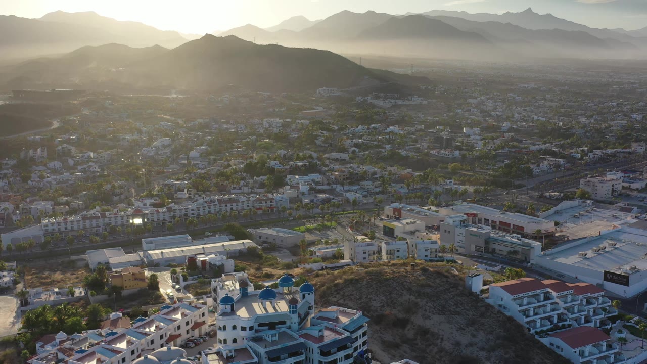 An aerial view of Los Cabos places a dry ridge and blue-domed white building in the foreground, with roads, rooftops, palms, and hazy mountains beyond, with no visible people.