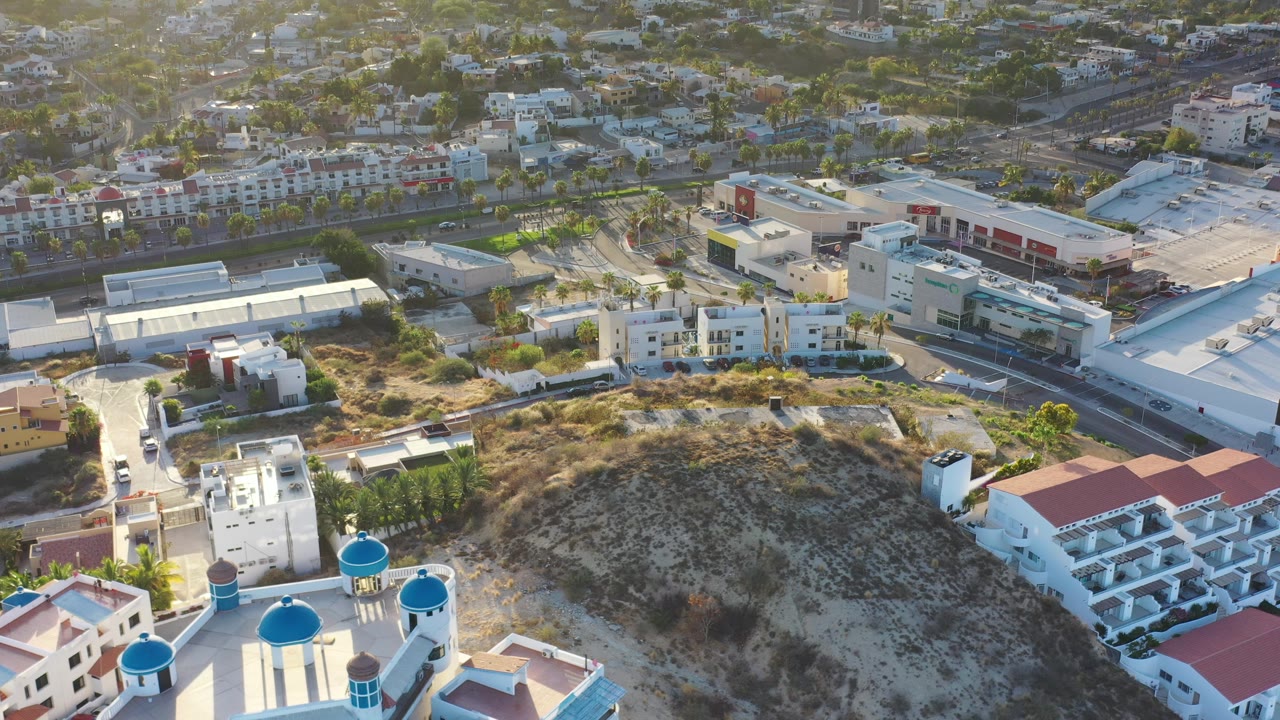 Los Cabos aerial frame showing the hill centered below the camera, blue-domed white architecture near the lower edge, red-roofed buildings to the side, and a mix of homes and commercial-looking structures beyond, with no visible people.