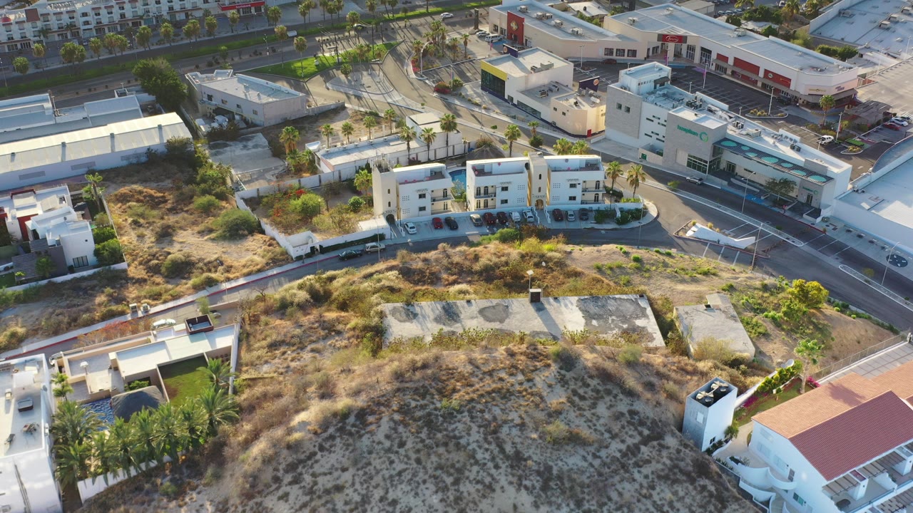 Opening aerial frame over Los Cabos, Mexico, showing white low-rise buildings beside curving roads, dry scrub lots, palms, and parked cars with no visible people.