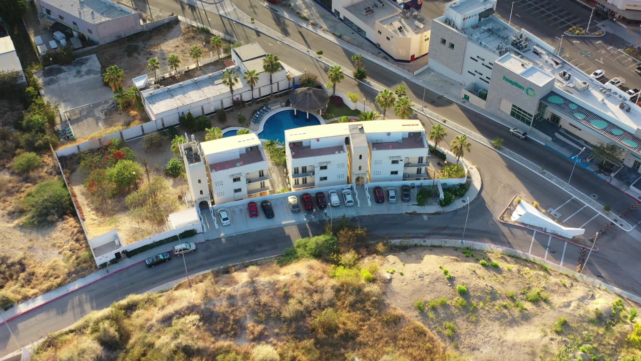 Aerial frame over Los Cabos residences with the apartment roofs, parking row, curved road, palm-lined pool area, and a larger building across the street, with no visible people.