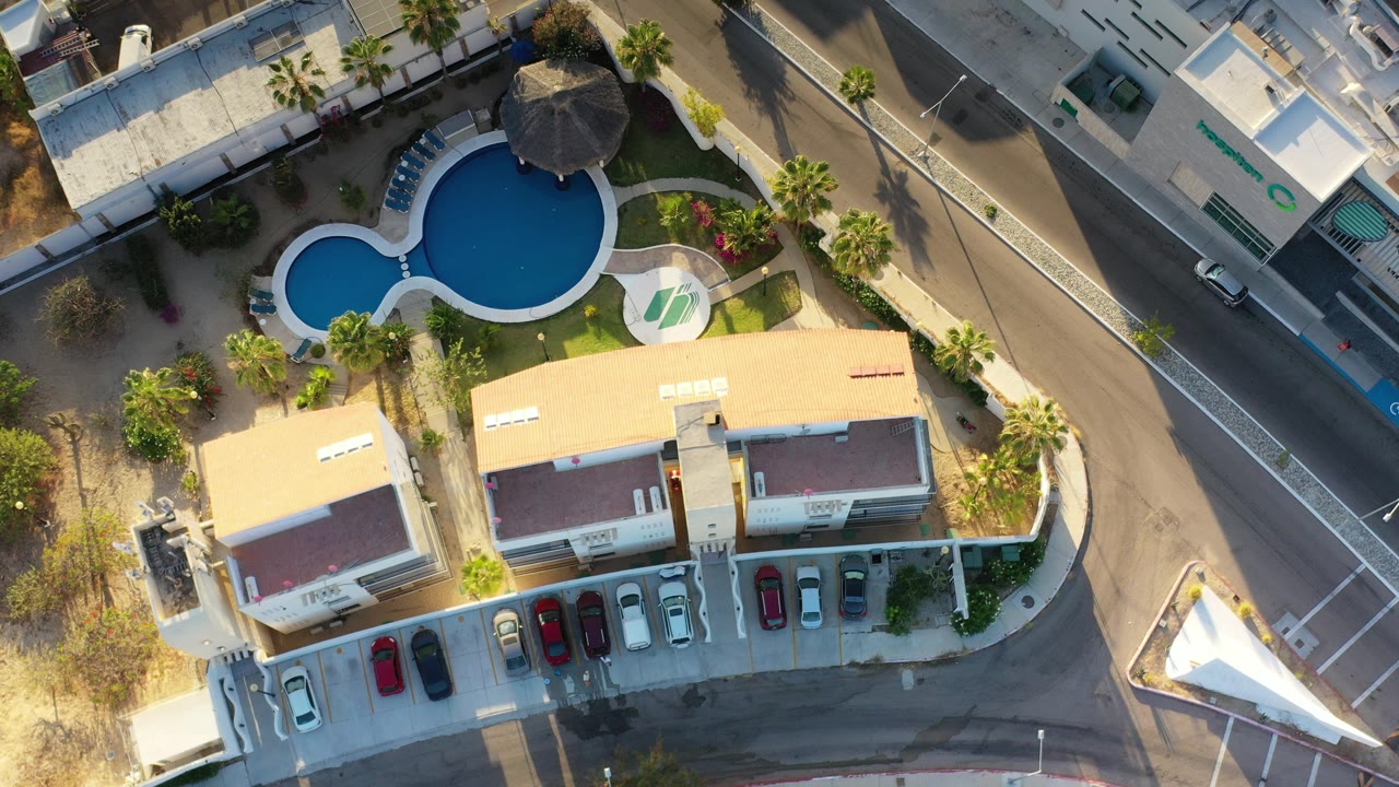 Overhead view of a hotel, blue swimming pool, road, palms, and parked cars.