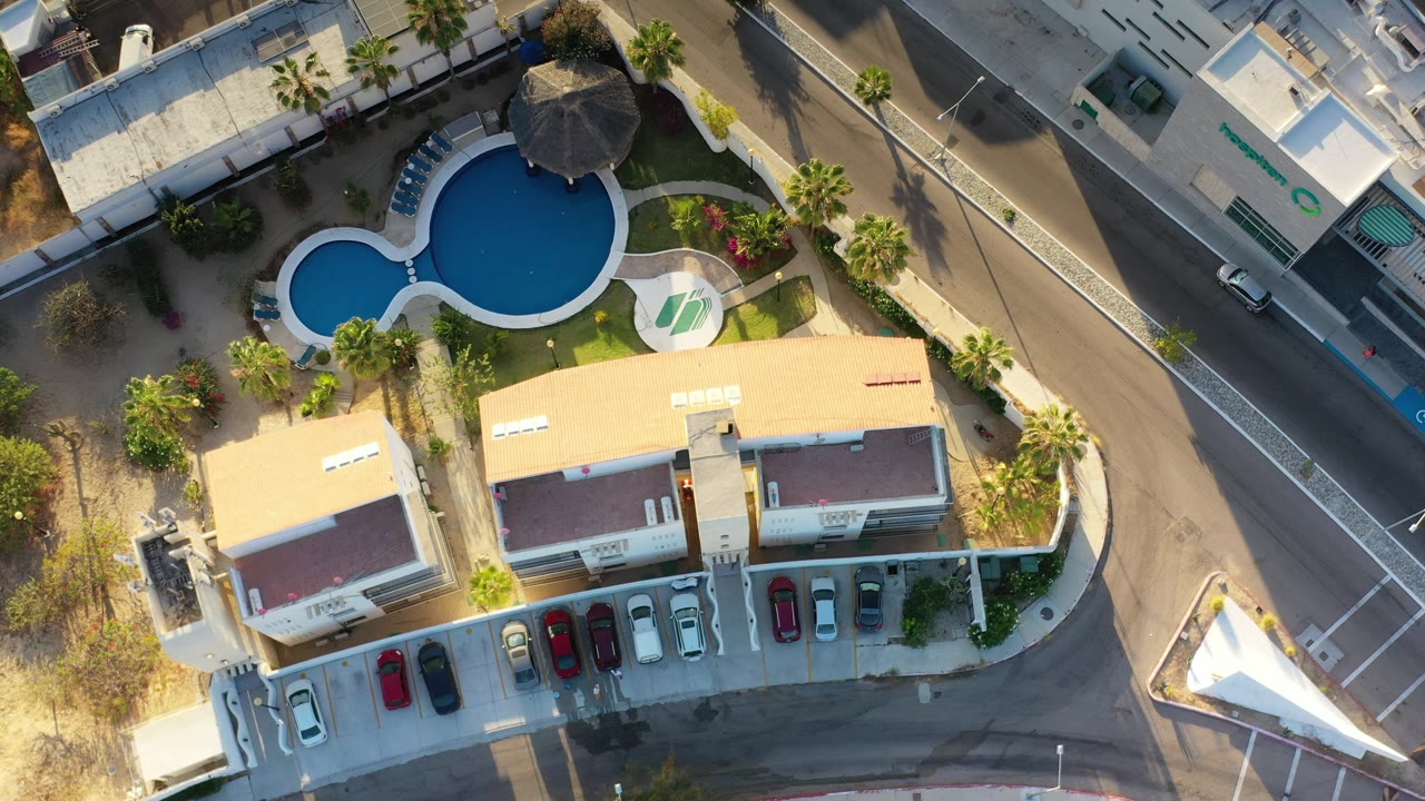 Overhead Los Cabos frame with the blue pool centered near the upper left, the hotel roof and parking lot below it, a road to the right, and no visible people.
