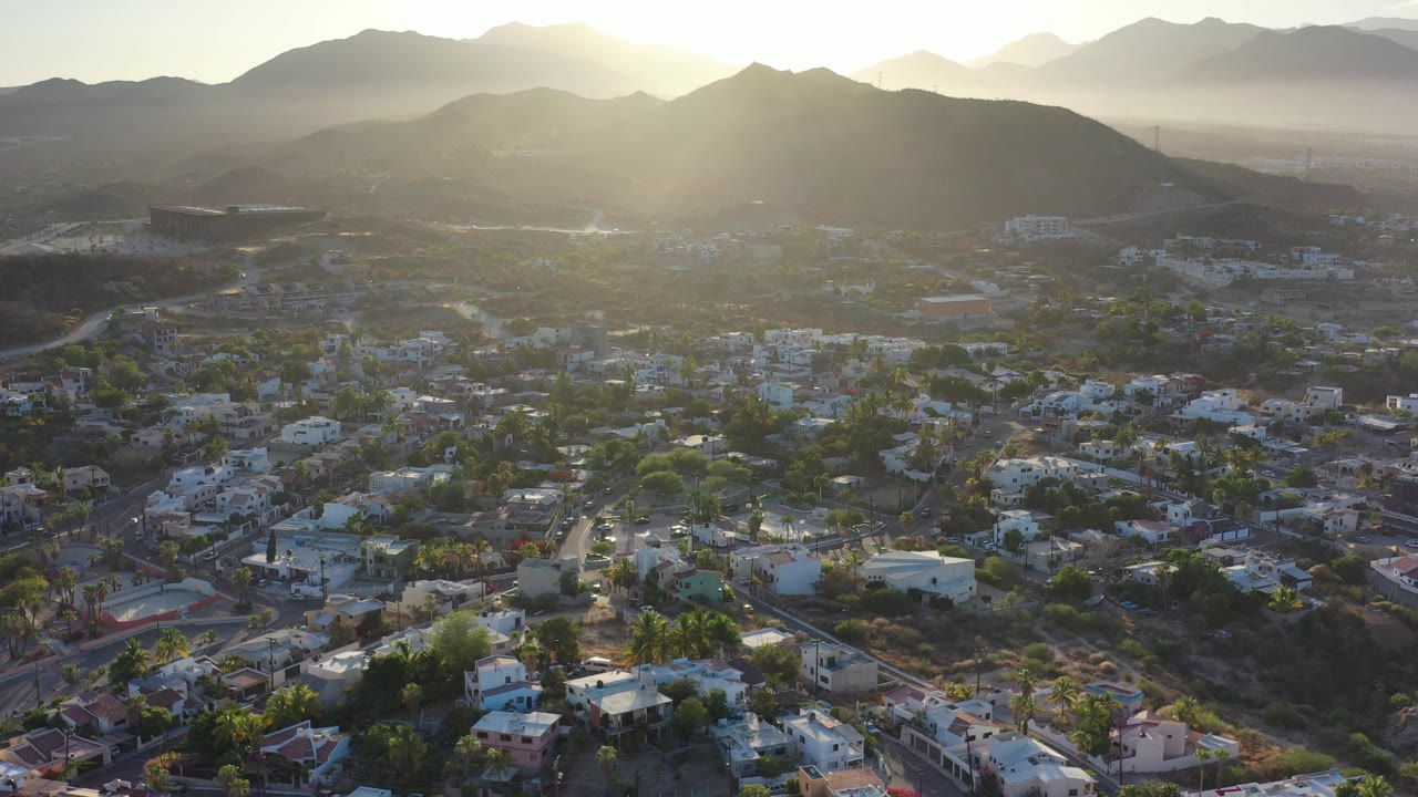 High aerial frame over Los Cabos, Mexico, showing low buildings, roads, palms, dry hills, and a bright sun flare above the mountain ridge with no visible people.