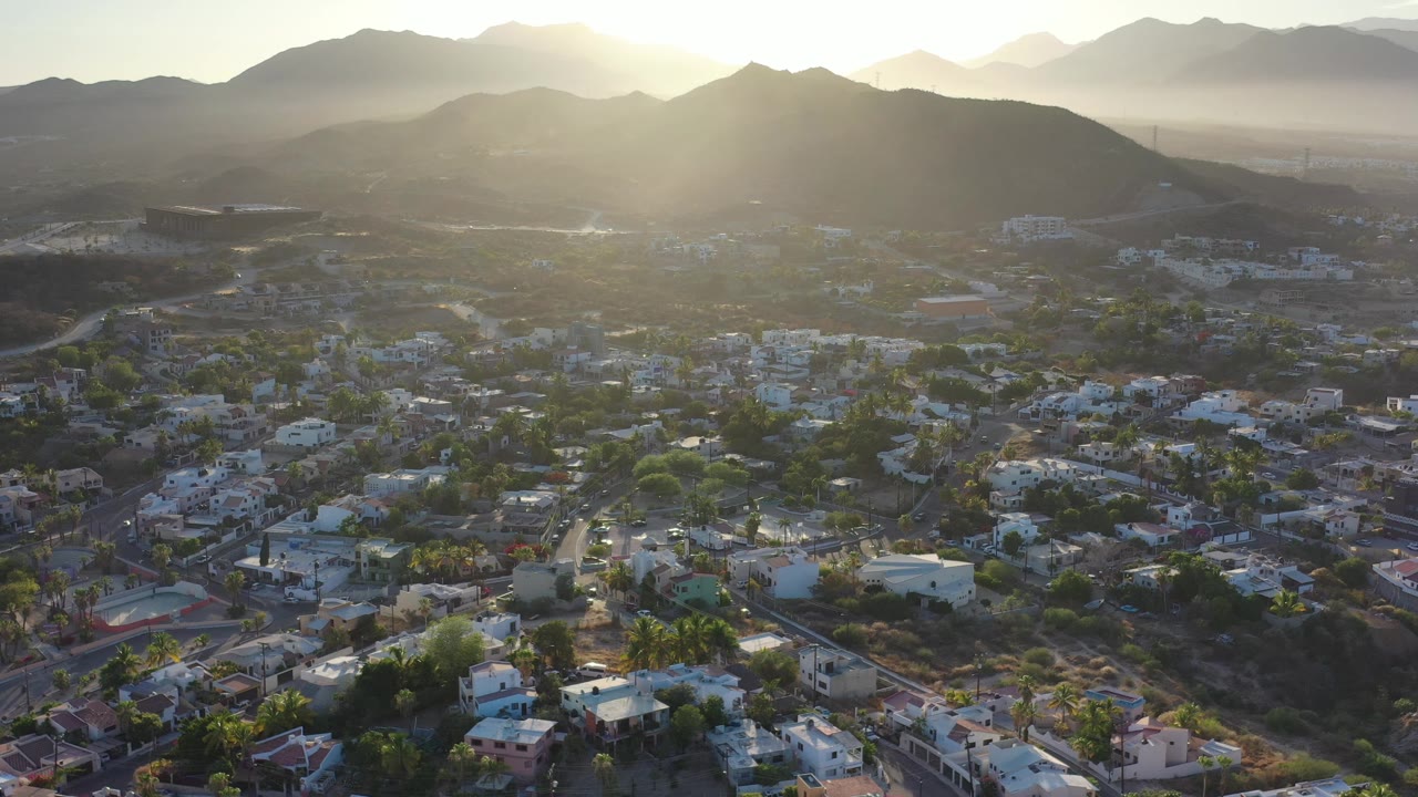 Wide drone view of rooftops, hills, and backlit mountains.
