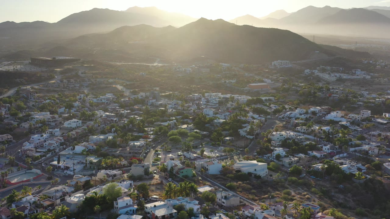 Opening aerial frame over Los Cabos, Mexico, showing clustered low-rise buildings, roads, palms, dry hills, hazy mountains, bright sun glare, and no visible people.
