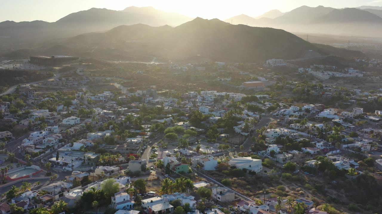 Aerial view across Los Cabos rooftops and greenery toward a dark mountain ridge, with the sun high near the horizon and no visible people.