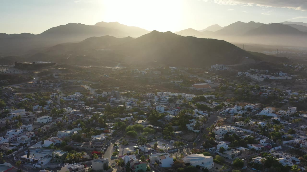 Los Cabos rooftops, palms, and roads sit in shadow below a central mountain ridge, with the sun flaring over distant hills and no visible people.