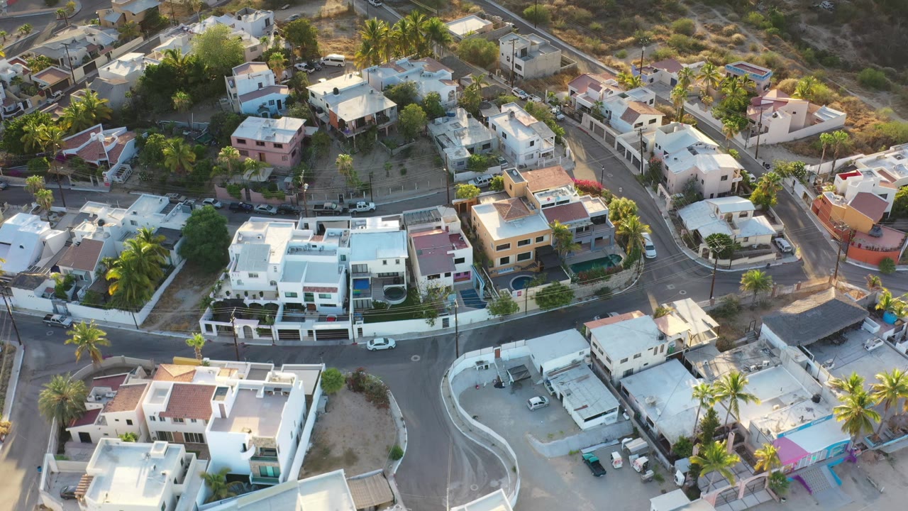 Los Cabos drone frame centered on a curved road bend, white and pastel homes, palm trees, parked vehicles, and dry open lots, with no visible people.