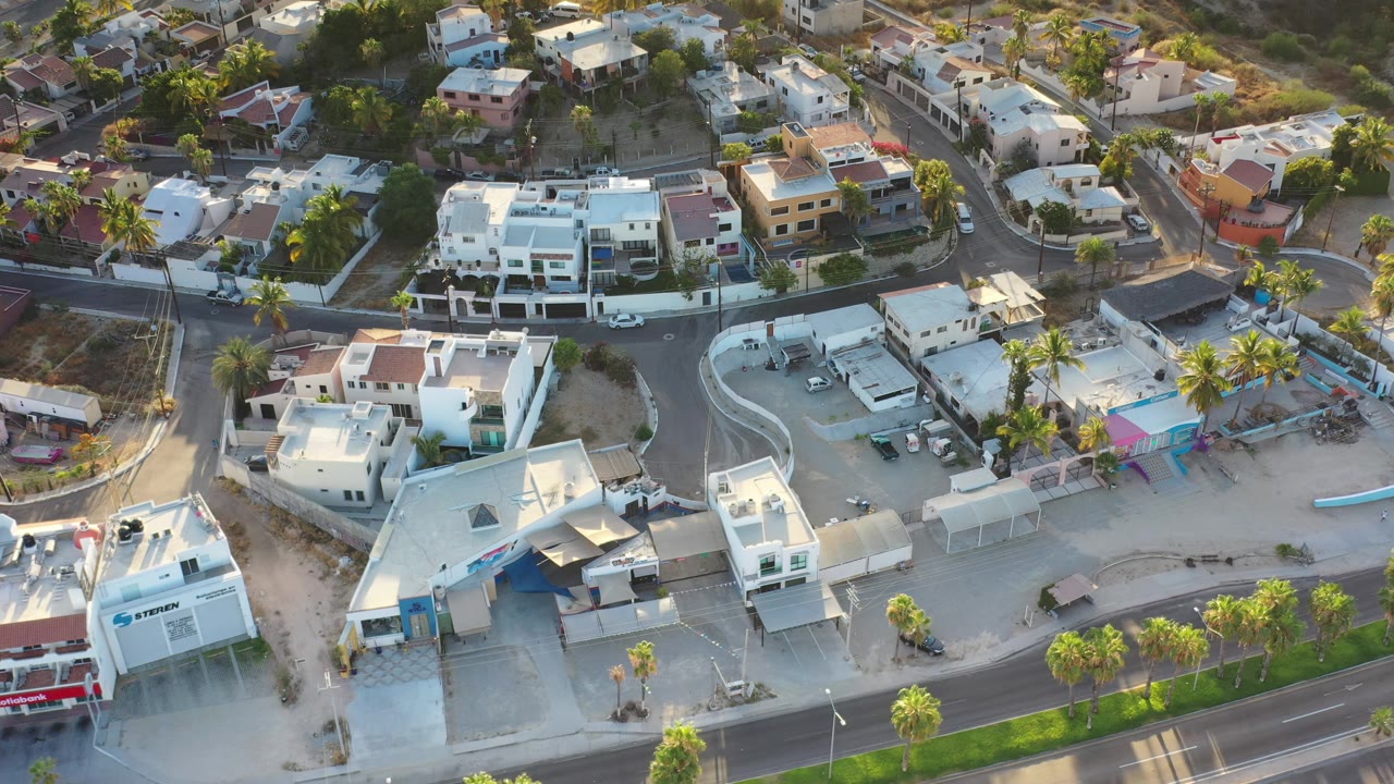 Early aerial frame in Los Cabos, Mexico, with a curved road, light-colored buildings, palms, parked vehicles, dry hillside homes, and no visible people.