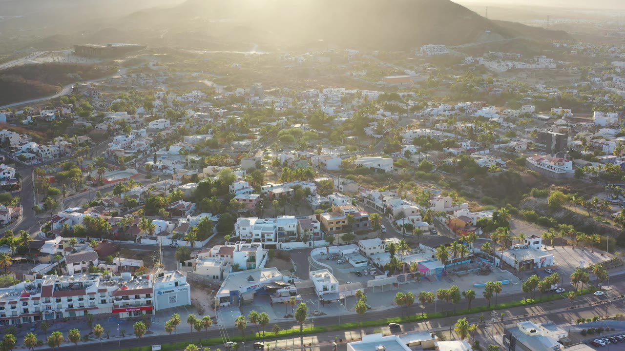 Los Cabos aerial frame showing a spread of white buildings, neighborhood roads, palm-lined streets, and a shadowed mountain slope behind them, with no visible people.