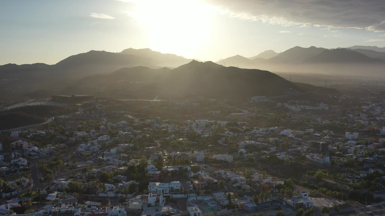 Opening frame of Los Cabos, Mexico, with bright sun above layered mountain ridges and a low-rise city in shadow below, no visible people.