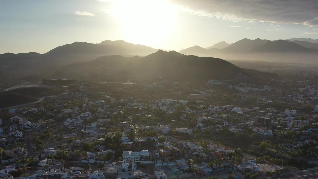 Los Cabos aerial view one second in, with the sun washing the upper sky and silhouetted mountains behind a dense low-rise city, no visible people.