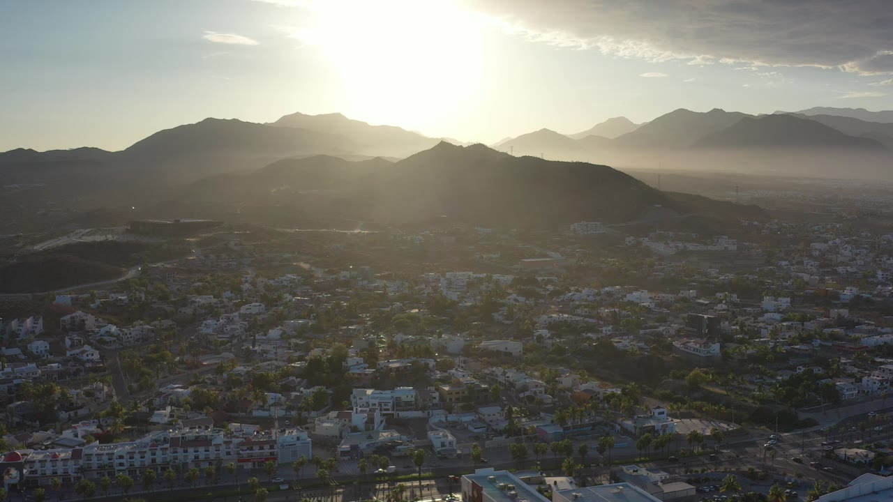Early frame of a Los Cabos sunrise with the sun high above dark mountain ridges, haze over the city, rooftops at the bottom edge, and no visible people.