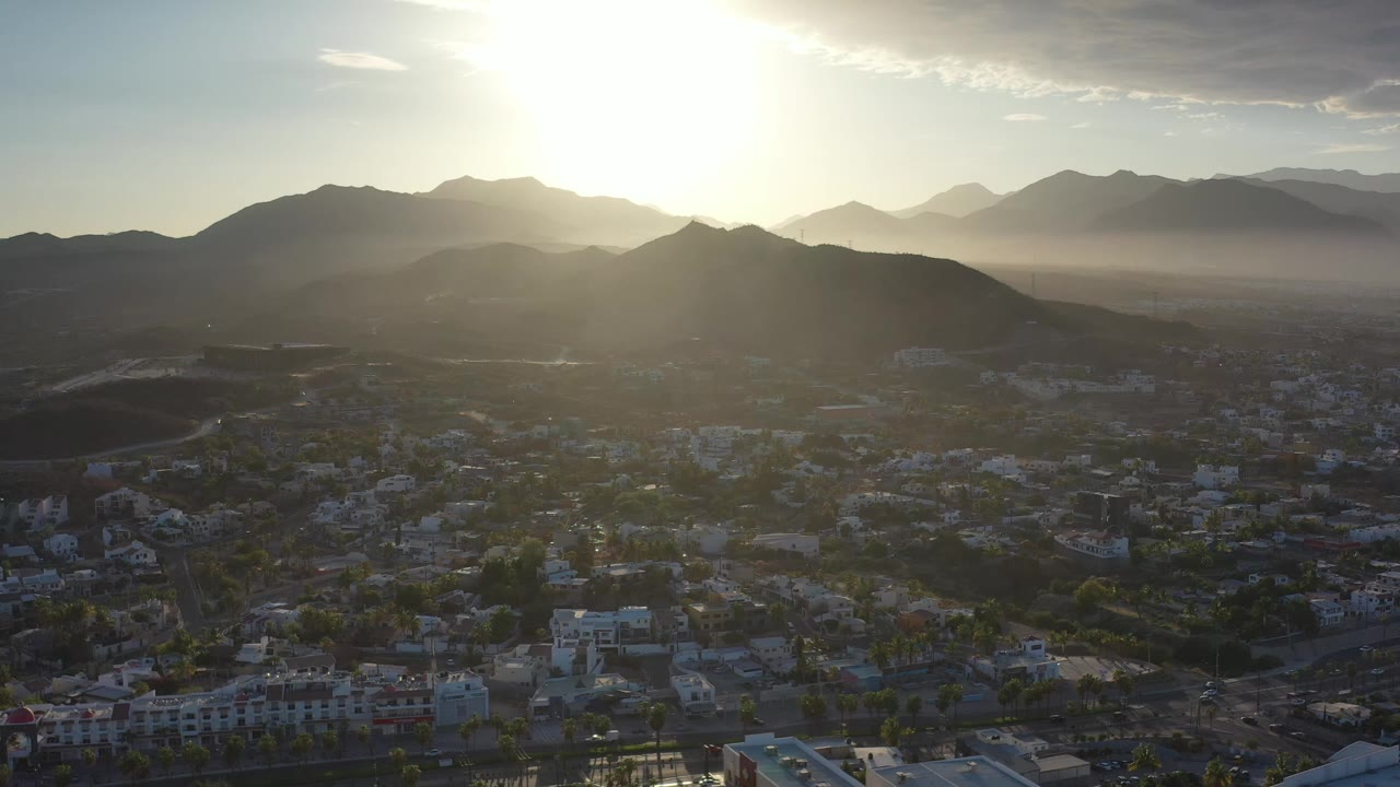 Los Cabos city blocks sit beneath layered mountain silhouettes as the bright sunrise glare spreads across the upper center of the frame, with no visible people.