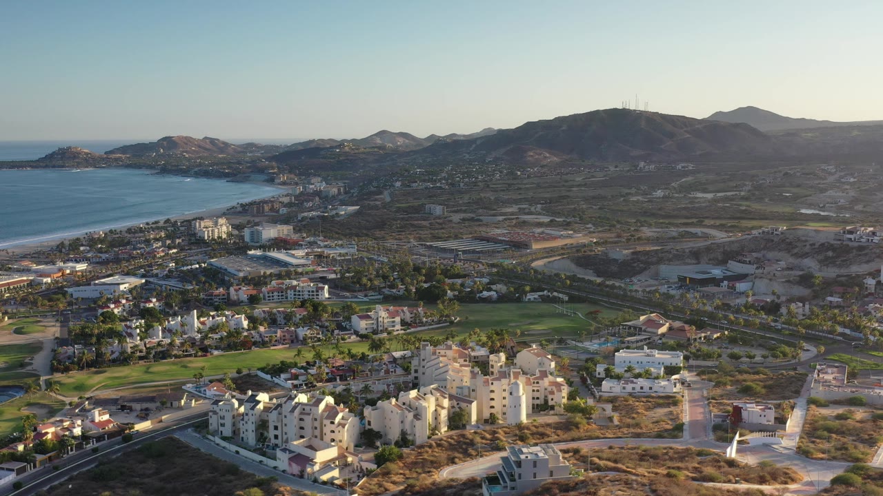 Wide aerial frame of Los Cabos, Mexico showing a blue shoreline at left, apartment-like buildings at bottom, green course areas through the center, and low mountains beyond, with no visible people.