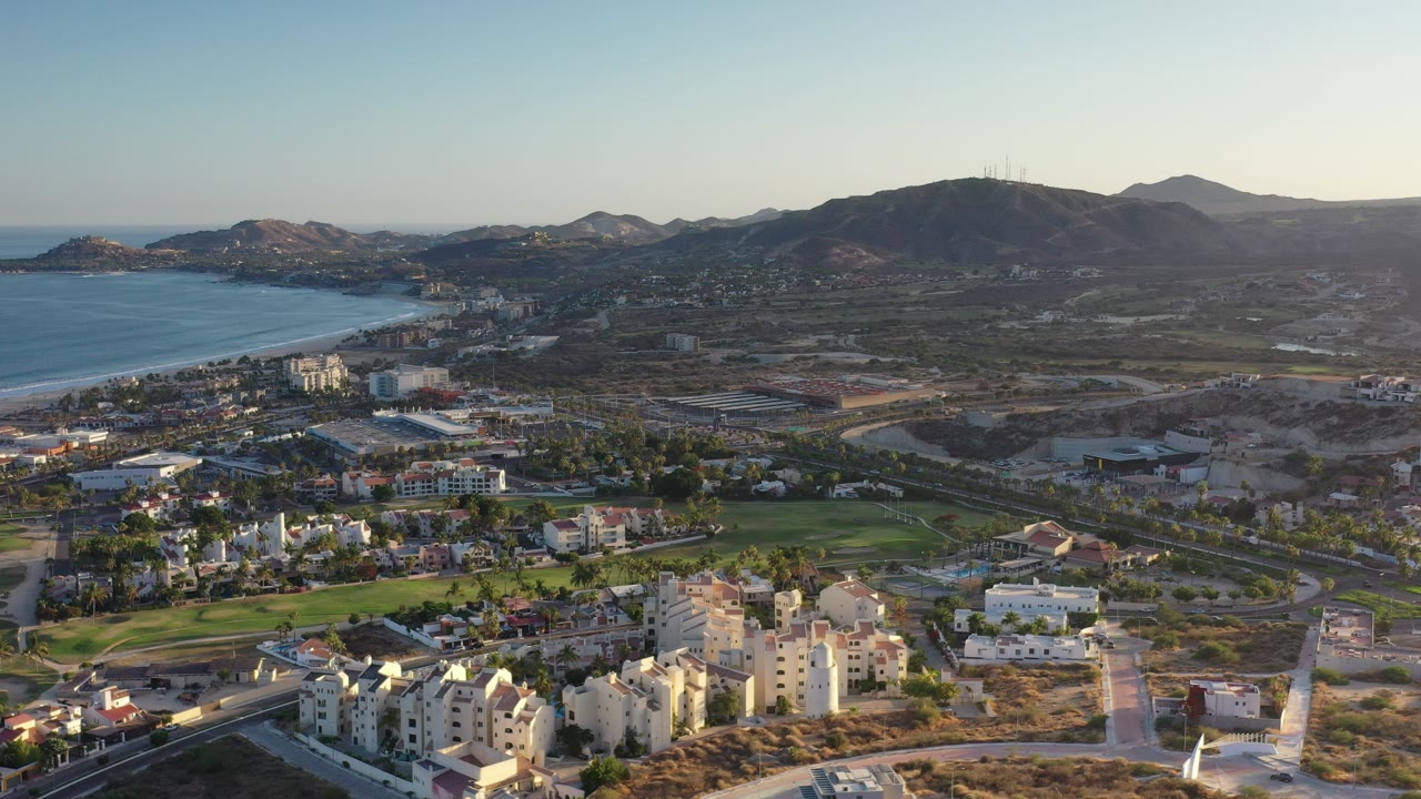 A wide aerial view over Los Cabos, Mexico shows low-rise buildings and roads in front, green fields near the center, a blue bay at left, and mountains under a clear sky, with no visible people.