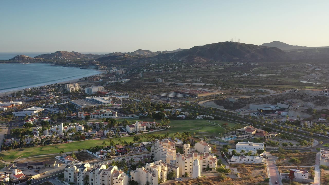 High aerial view over Los Cabos, Mexico, with blue ocean and beach on the left, resort buildings and golf greens below, dry land and mountains beyond, and no visible people.