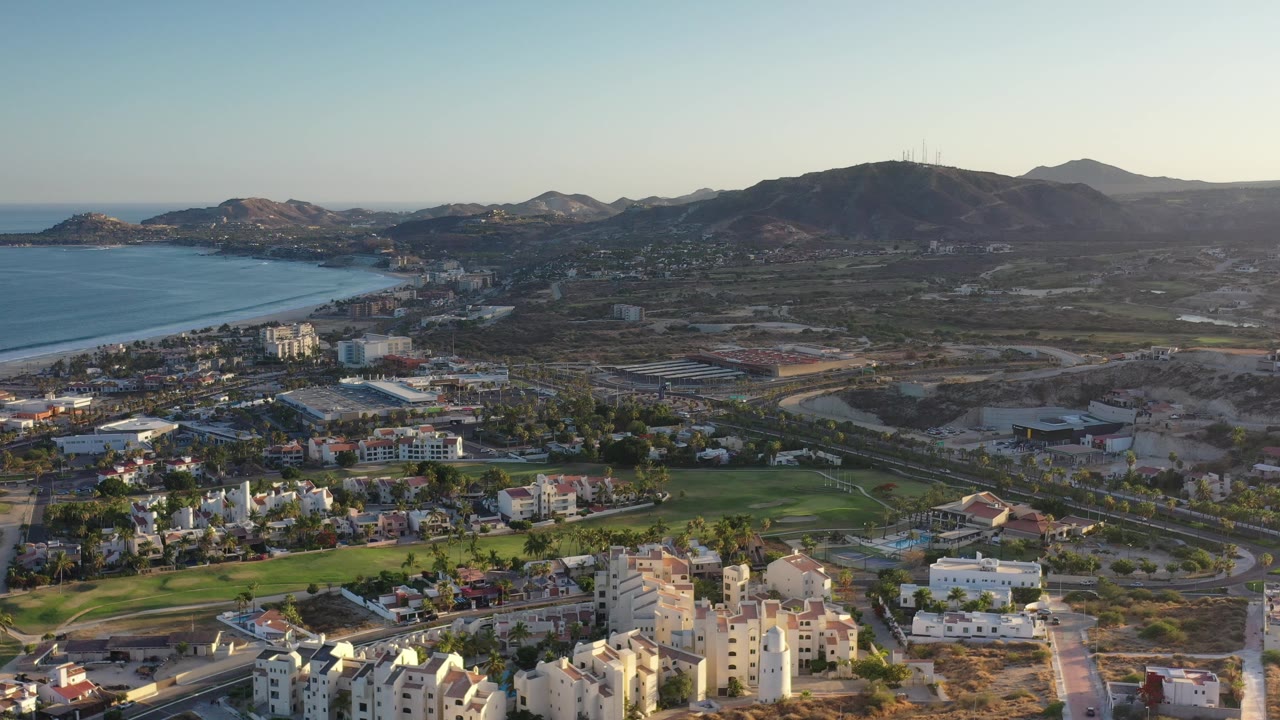Drone-style view of Los Cabos coastline, hotels, roads, golf course, and hills.