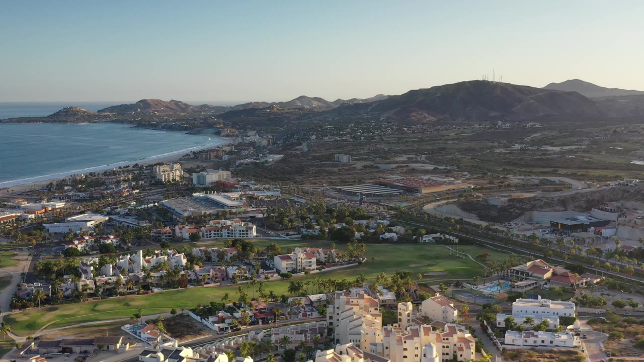 Aerial frame one second into the clip shows Los Cabos, Mexico, with blue water along the left edge, clustered buildings below, a green course-like area, distant hills, and no visible people.
