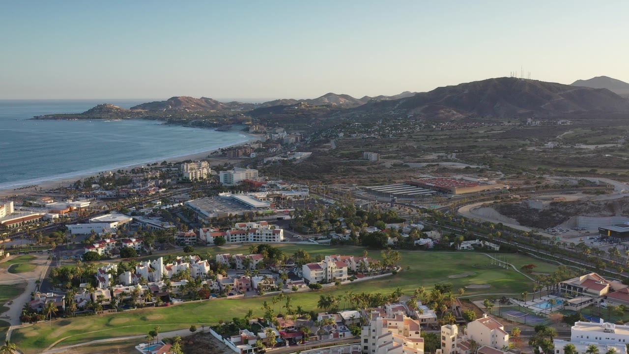 Opening aerial frame over Los Cabos with ocean and a curving beach on the left, resort and residential buildings below, dry hills beyond, and no visible people.