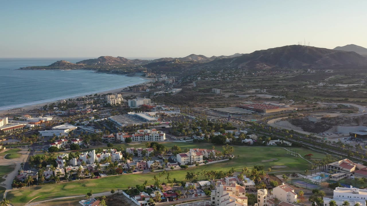 Los Cabos aerial view one second in, showing the surf line, beachside town, clustered homes, green open space, and mountain ridge under soft light with no visible people.