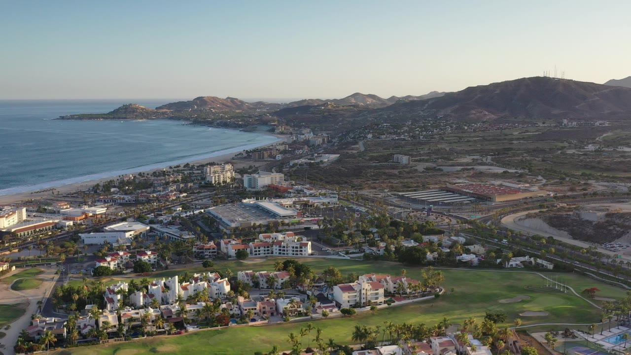 Wide aerial view over Los Cabos, Mexico, with the ocean and curving beach on the left, low-rise city buildings and golf-course greenery below, hills in the distance, and no visible people.