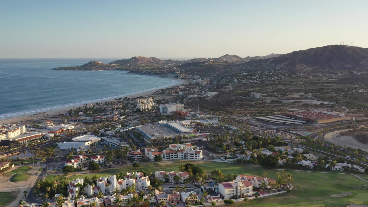 Opening aerial frame over Los Cabos, Mexico, with blue sea and a long beach at left, low-rise buildings and green landscaping below, hills on the horizon, and no visible people.