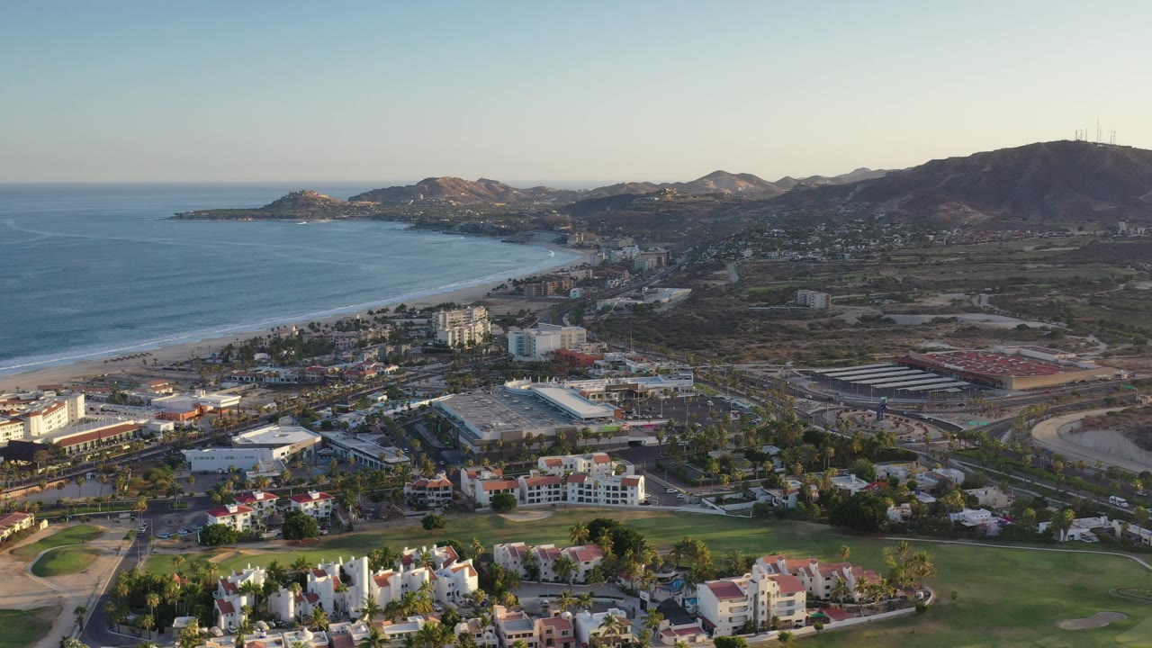 Elevated shoreline view with sea, beach, clustered buildings, green foreground, and hills.