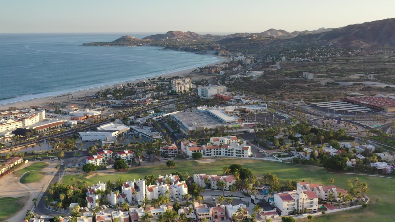 Aerial view of a Los Cabos bay, beach, buildings, golf course, and hills.