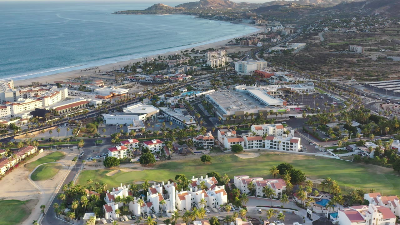 Aerial frame in Los Cabos, Mexico showing the beach and blue water above a cluster of white buildings, paved roads, palm rows, golf turf, and private homes, with no visible people.