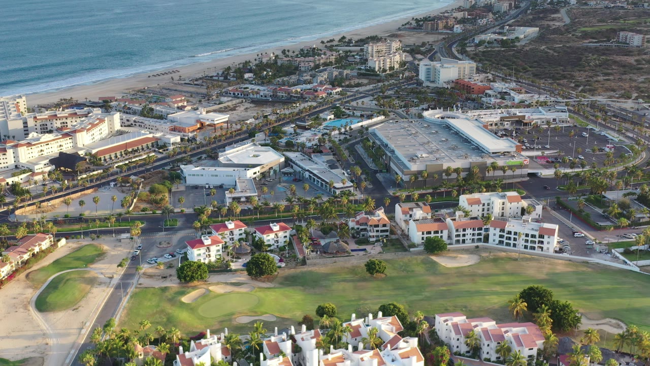 Los Cabos aerial view one second in, showing surf along the beach, a diagonal road network, hotel-like buildings, parked and moving vehicles, and a golf green below, no visible people.
