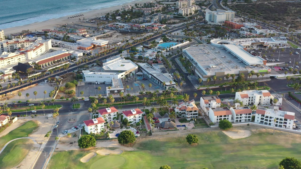 The aerial view in Los Cabos, Mexico holds over red-roofed residences, a broad road with cars, large flat-roof buildings, beach sand, and blue ocean, with no visible people.