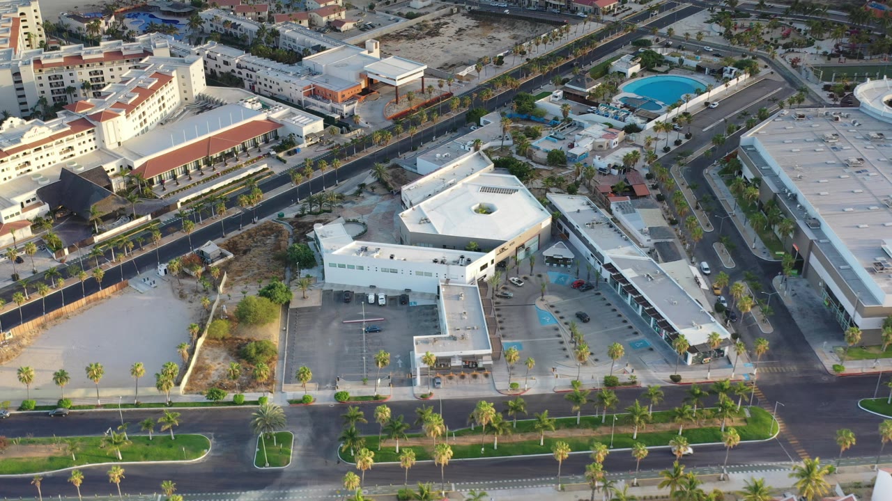 Oblique aerial frame in Los Cabos, Mexico, with a white angular building centered, a dark road along the lower edge, parked cars, palms, and no visible people.