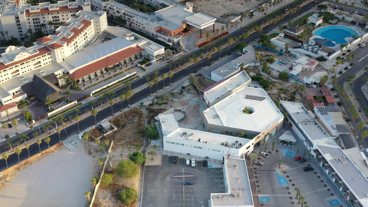 Aerial view over Los Cabos, Mexico, with a palm-lined road crossing between a white angular building, parking areas, dry lots, and no visible people.