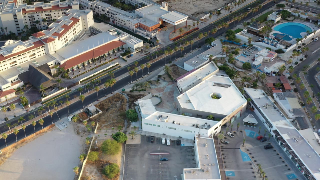 Aerial Los Cabos frame showing the road, rows of palms, a white rooftop in the foreground, apartment-style buildings beyond, a blue pool, and no visible people.