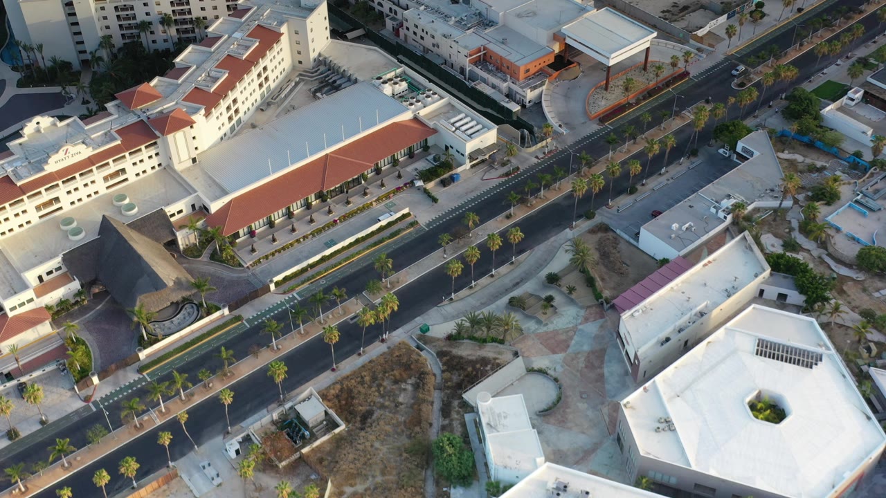 A second aerial view in Los Cabos looks across the divided avenue, small cars, palm trees, white rooftops, and empty sidewalks with no visible people.