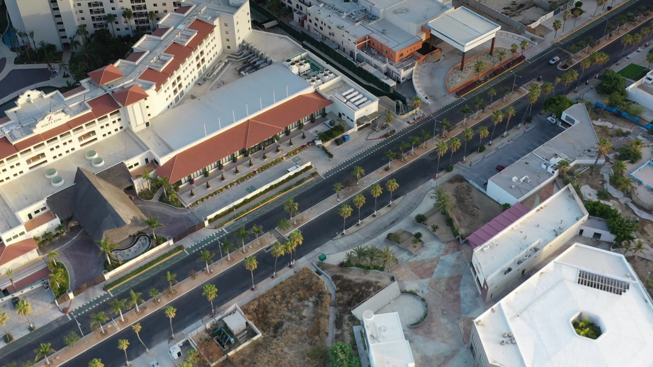 Aerial city view with red-roofed buildings, a road, palms, and paved plazas.