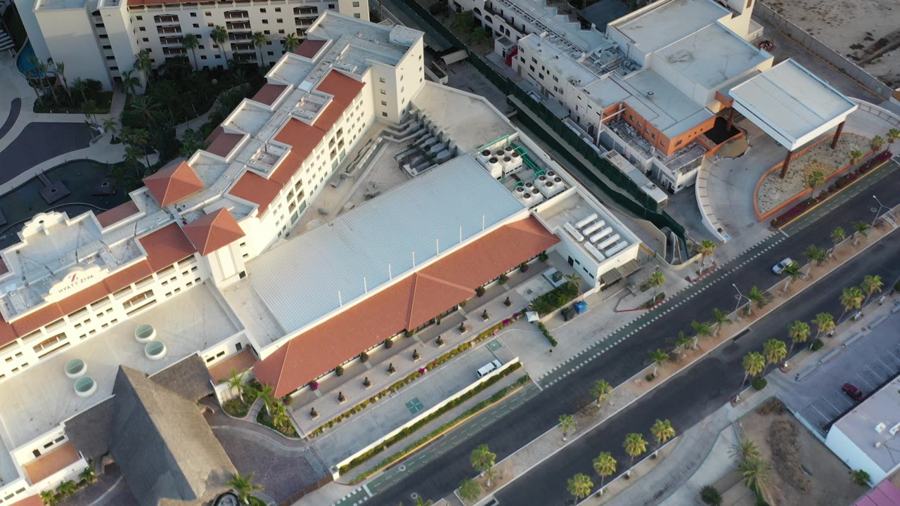 Opening aerial frame over Los Cabos, Mexico, showing cream multi-story buildings, red tile roof sections, palm-lined streets, and no visible people.