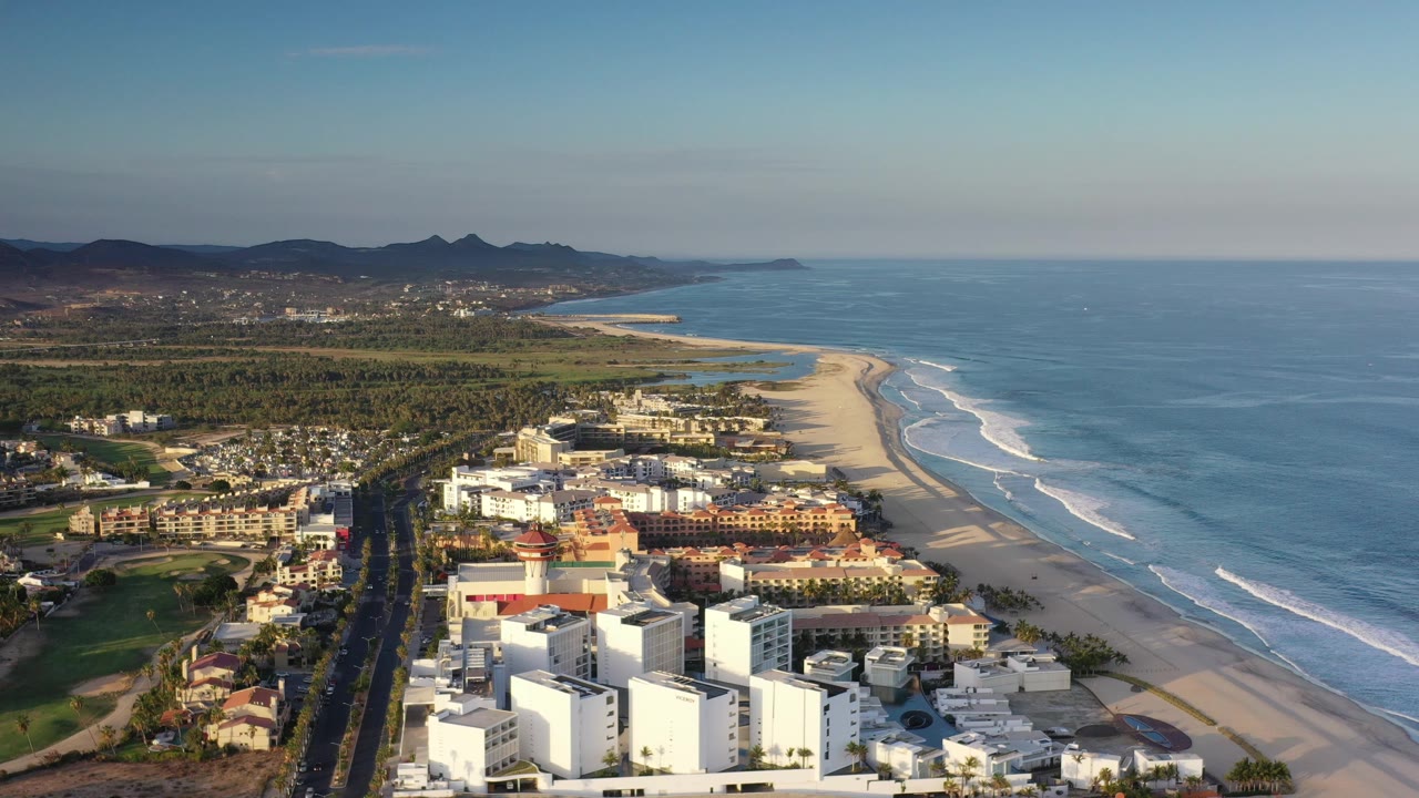 Early aerial frame over Los Cabos, Mexico showing white beachfront buildings in the foreground, a long sandy beach, blue surf to the right, inland greenery, distant hills, and no visible people.
