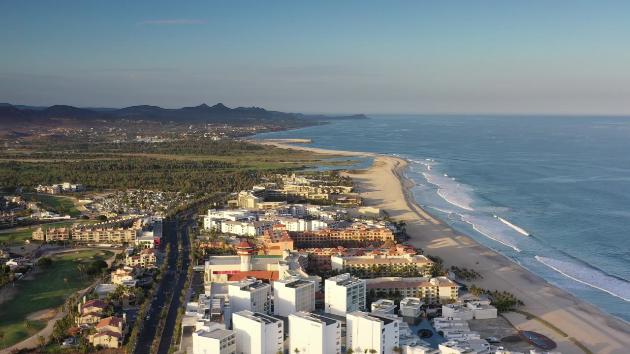A high aerial frame over Los Cabos shows white beachfront buildings in the foreground, a long sandy beach, blue ocean to the right, distant hills, and no visible people.