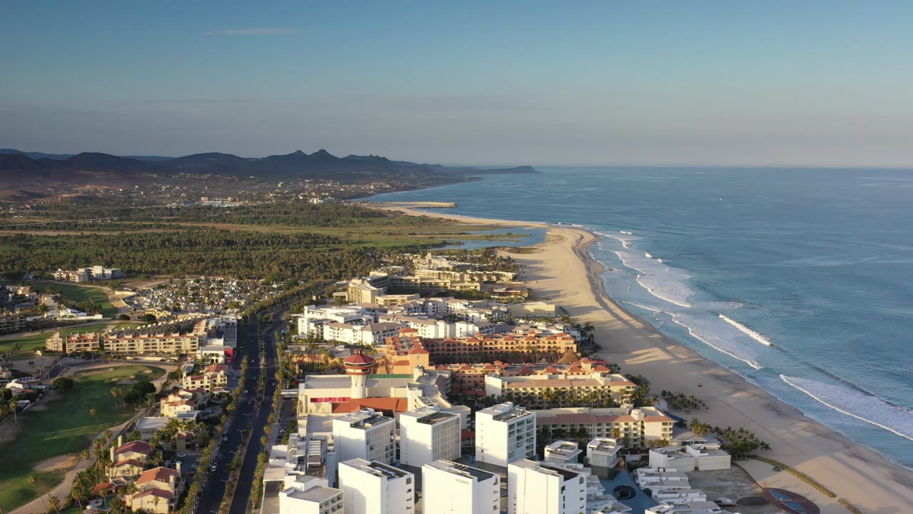 Drone-height view of a resort coastline, open beach, surf, ocean, and inland hills.