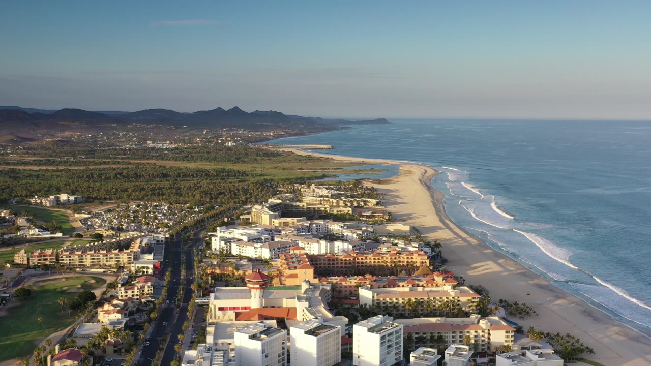 Early frame of Los Cabos, Mexico from above, with resort and city buildings in the foreground, a sandy beach and white surf to the right, distant hills inland, and no visible people.