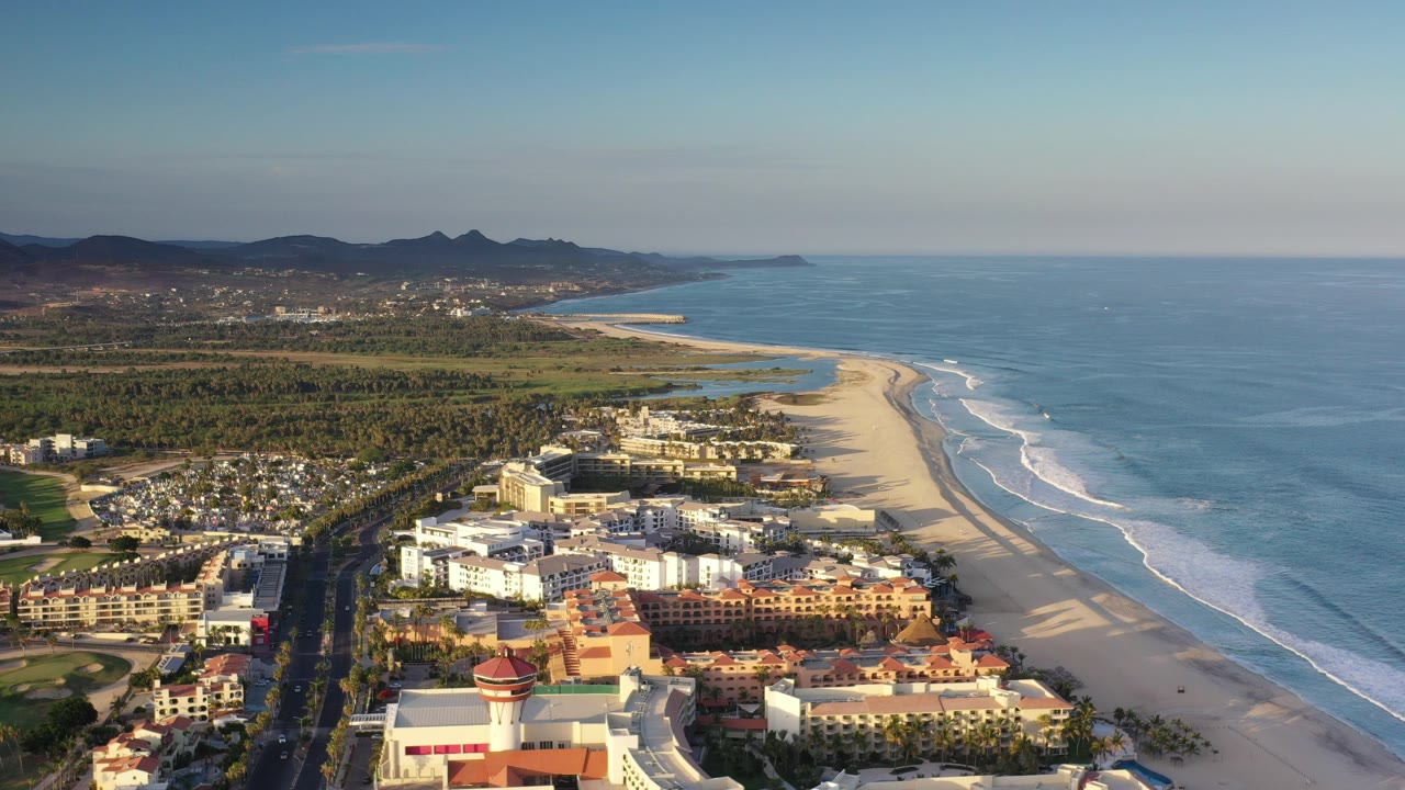 Aerial view in Los Cabos, Mexico, showing low-rise buildings near a broad sandy beach, blue ocean surf to the right, distant hills, and no visible people.