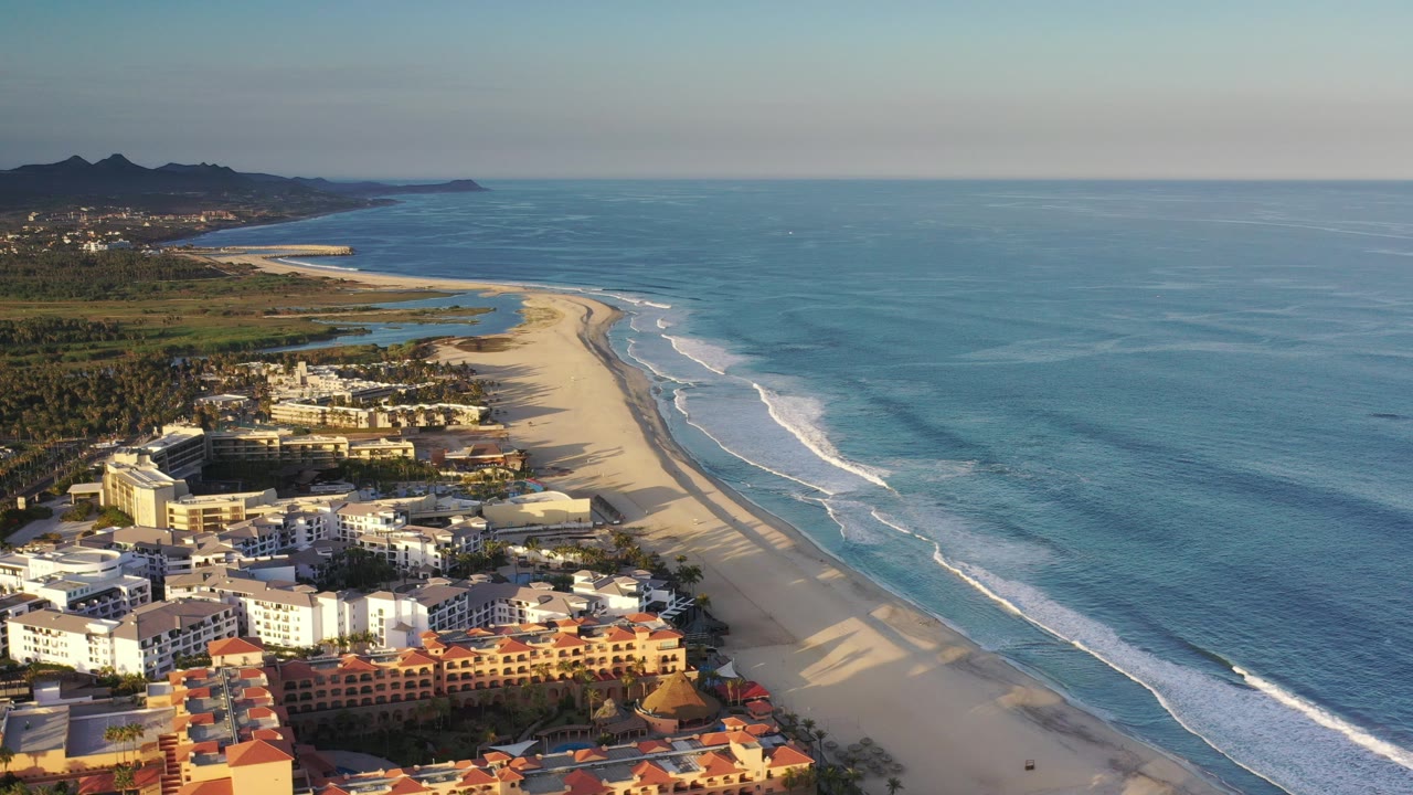 Opening aerial frame over the Los Cabos coastline, with beachfront buildings at lower left, a long pale beach, white surf lines, blue ocean, distant hills, and no visible people.
