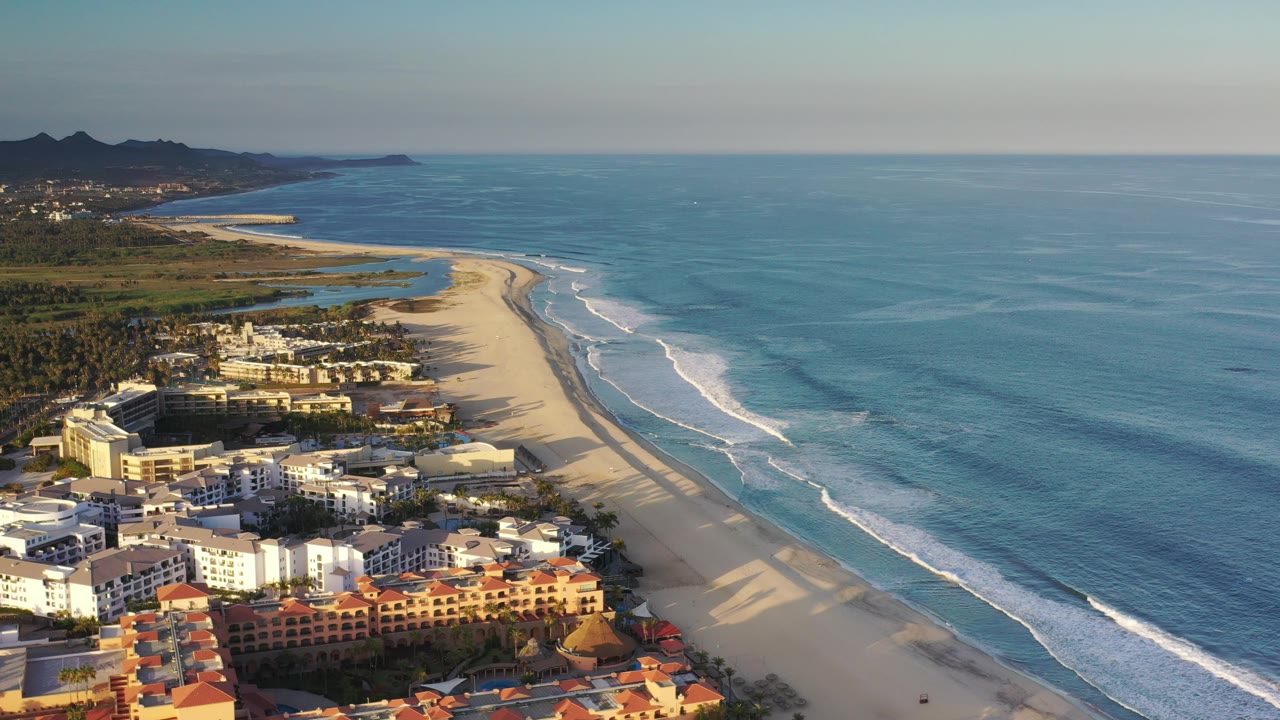 Elevated shoreline view with white surf bands beside beach hotels and greenery.