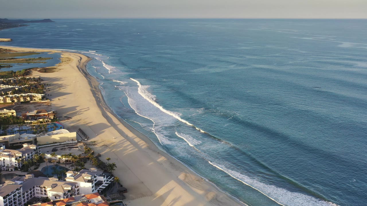 Aerial Los Cabos shoreline frame with the beach curving toward the top left, white breakers spreading along the waterline, and no visible people on the sand.