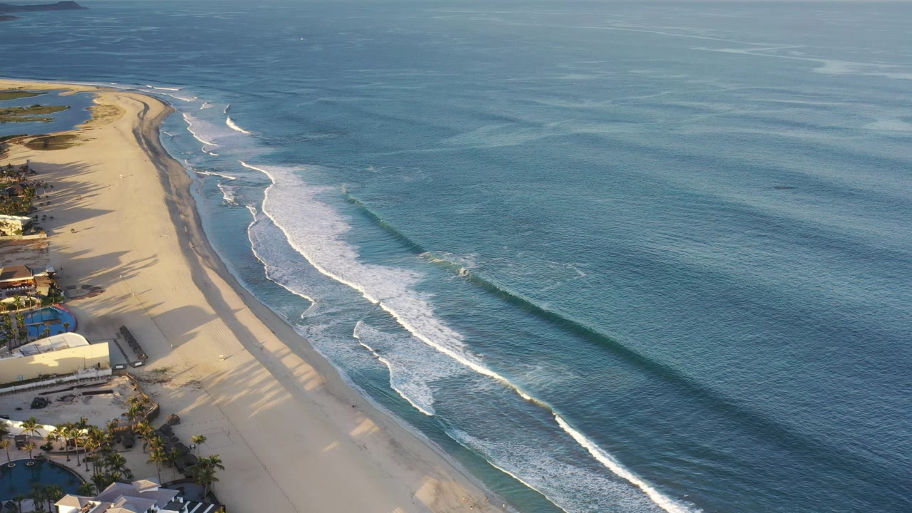 Aerial view of sandy coastline with white surf and blue ocean.